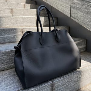 May include: A black handbag with a textured surface, featuring two rounded top handles and a horizontal seam across the front. The bag has a structured base and curved side panels, showcasing a solid black color throughout the exterior design.