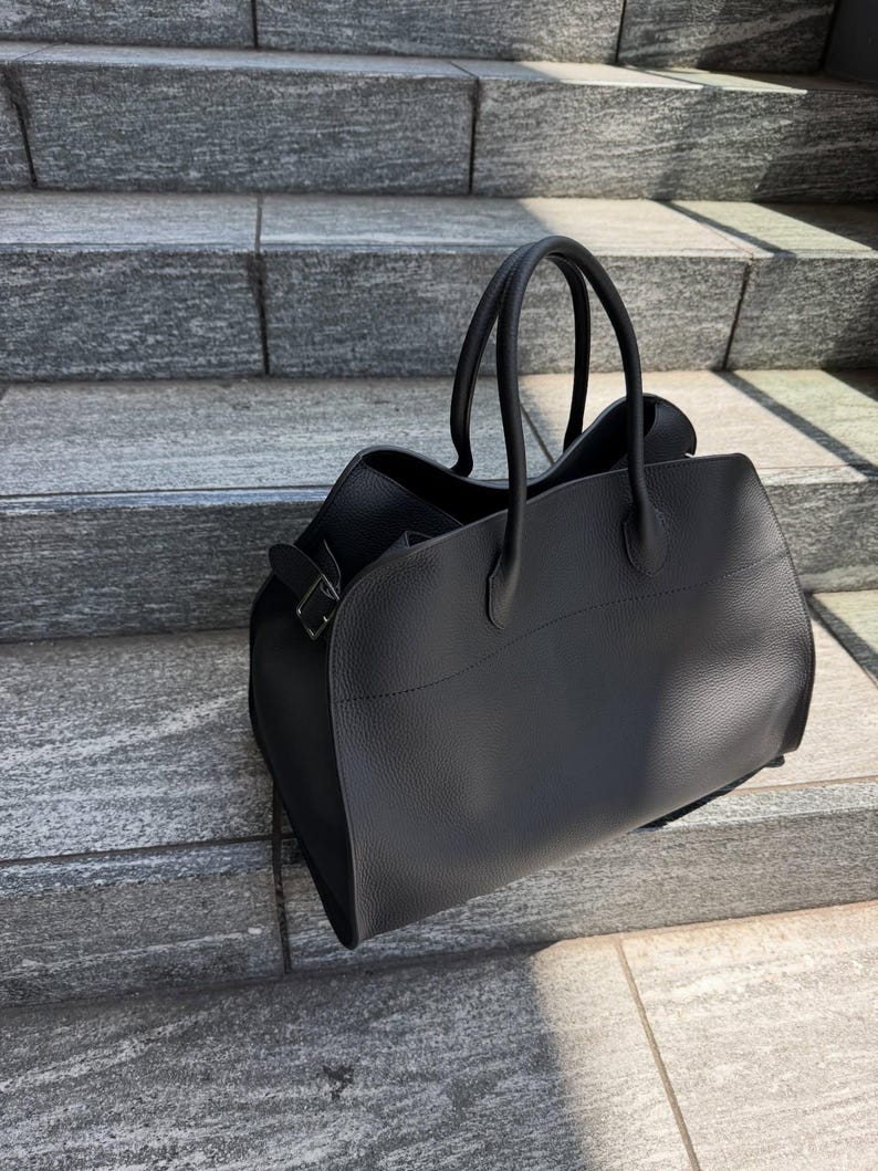 May include: A black handbag with a textured surface, featuring two rounded top handles and a side buckle detail. The bag has a structured base and a wide opening, showcasing a clean, solid color design with visible stitching along the top panel.