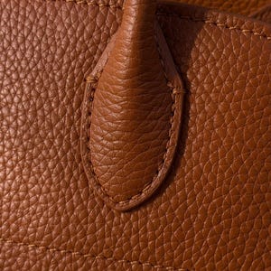 May include: A close-up view of a tan-colored bag featuring a textured, pebbled surface. The image highlights a sturdy handle attachment point, reinforced with matching tan stitching that follows the oval-shaped contour of the leather tab.