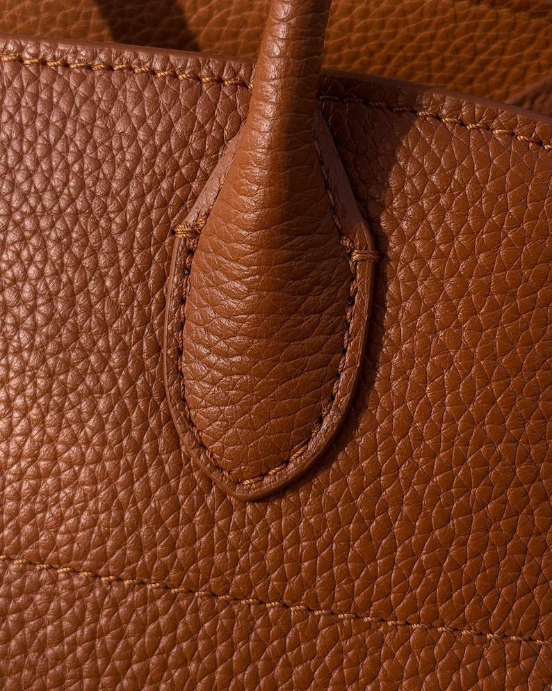 May include: A close-up view of a tan-colored bag featuring a textured, pebbled surface. The image highlights a sturdy handle attachment point, reinforced with matching tan stitching that follows the oval-shaped contour of the leather tab.