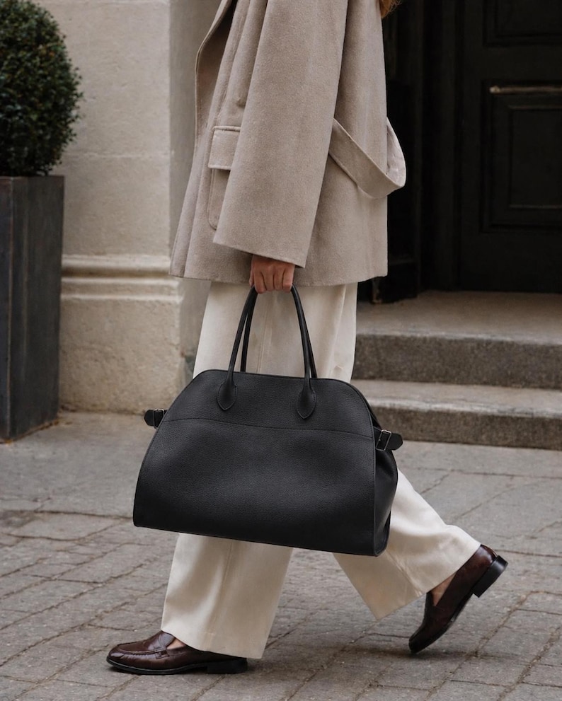 May include: A large black leather handbag with a rounded top silhouette, dual top handles, and side buckle straps. The bag features a textured finish and a spacious main compartment, providing a structured yet functional design for carrying daily essentials.