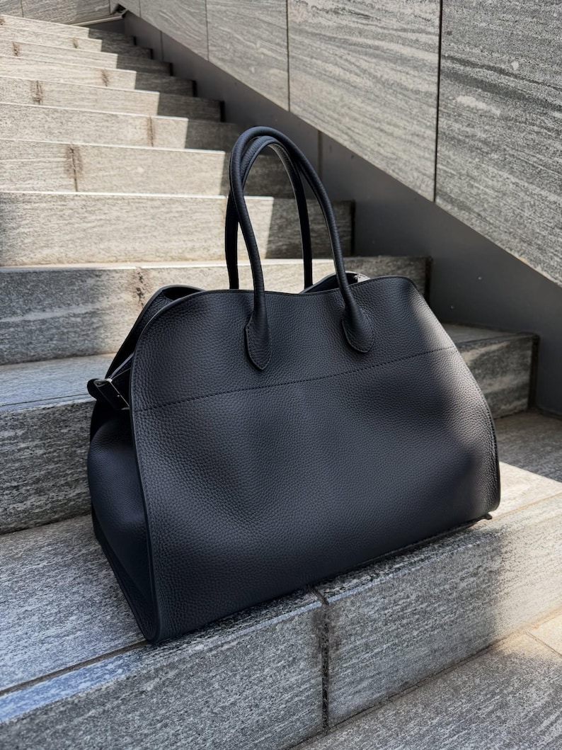 May include: A black handbag with a textured surface, featuring two rounded top handles and a horizontal seam across the front. The bag has a structured base and curved side panels, showcasing a solid black color throughout the exterior design.