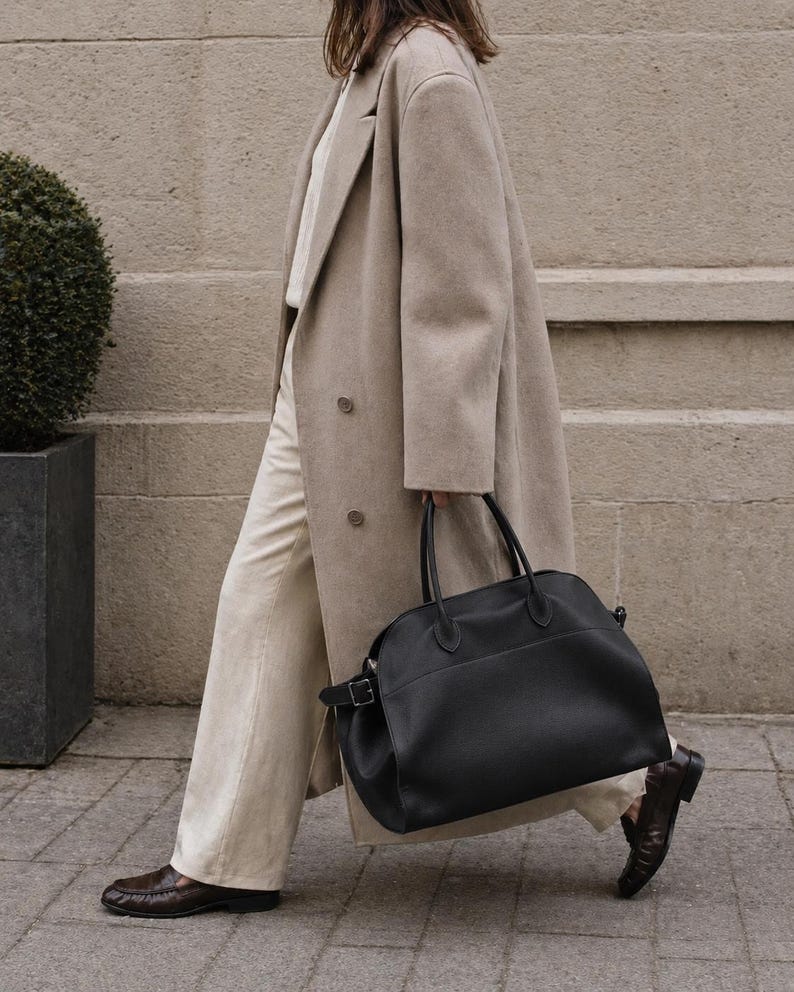 May include: A beige double-breasted wool coat worn over a white shirt and light-colored trousers, paired with dark brown leather loafers and a large black leather handbag featuring dual top handles and a side buckle detail.