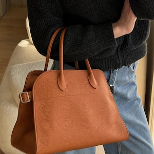 May include: A structured tan handbag with a pebbled texture, featuring two top handles and a silver-toned metal buckle detail on the side. The bag has a curved top silhouette and a spacious main compartment, crafted with clean lines and minimal hardware.