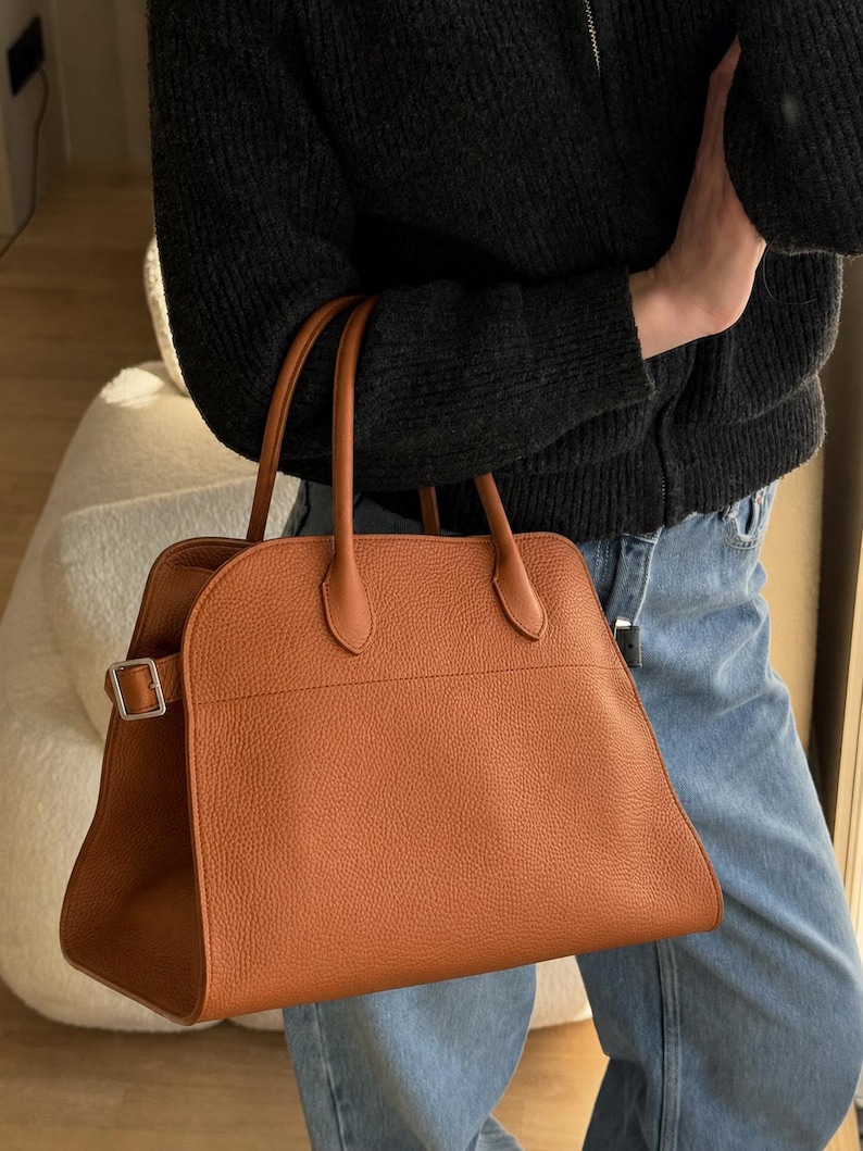 May include: A structured tan handbag with a pebbled texture, featuring two top handles and a silver-toned metal buckle detail on the side. The bag has a curved top silhouette and a spacious main compartment, crafted with clean lines and minimal hardware.