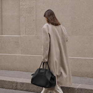 May include: A long beige coat worn over white wide-leg trousers and dark brown shoes, paired with a large black leather handbag featuring dual top handles and side buckle details, captured from a side profile view while walking.