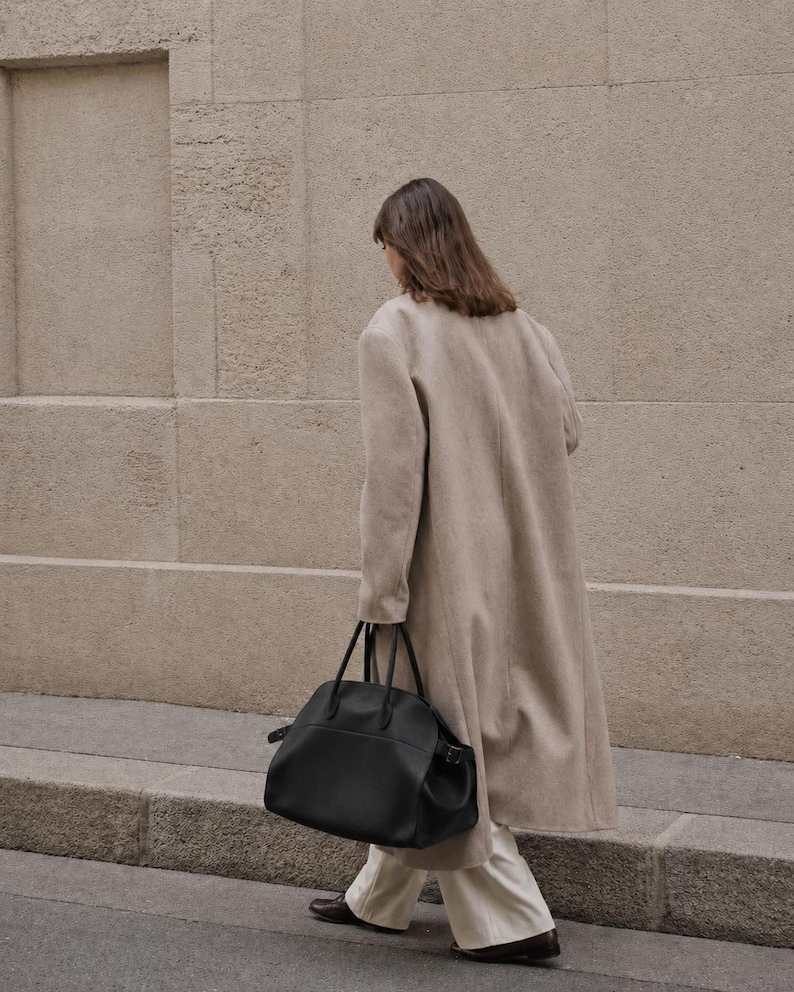 May include: A long beige coat worn over white wide-leg trousers and dark brown shoes, paired with a large black leather handbag featuring dual top handles and side buckle details, captured from a side profile view while walking.