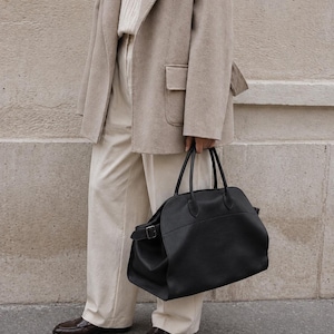 May include: A large black leather handbag with dual top handles and a side buckle strap, paired with a beige wool blazer, a cream ribbed sweater, cream trousers, and dark brown loafers. The handbag features a rounded silhouette and smooth texture.