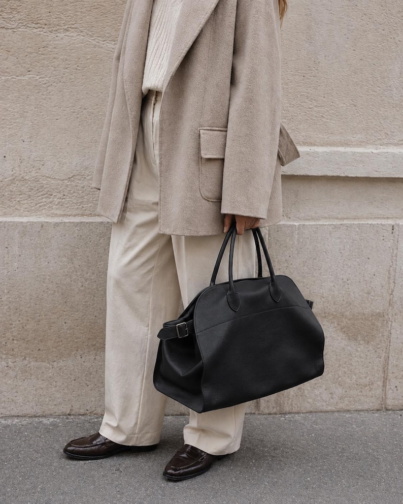 May include: A large black leather handbag with dual top handles and a side buckle strap, paired with a beige wool blazer, a cream ribbed sweater, cream trousers, and dark brown loafers. The handbag features a rounded silhouette and smooth texture.