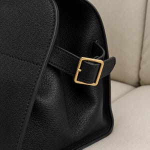 May include: A black textured leather handbag featuring a side strap with a polished gold-tone metal buckle. The bag has a structured design with visible stitching along the edges and a smooth, rounded side profile with a matte finish.