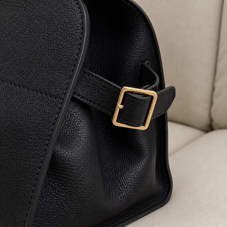 May include: A black textured leather handbag featuring a side strap with a polished gold-tone metal buckle. The bag has a structured design with visible stitching along the edges and a smooth, rounded side profile with a matte finish.