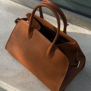 May include: A medium-sized, structured handbag in a rich cognac brown color with a distinct pebbled texture. The bag features two rounded top handles, a wide open-top design, and silver-toned metal hardware accents on the side attachments.