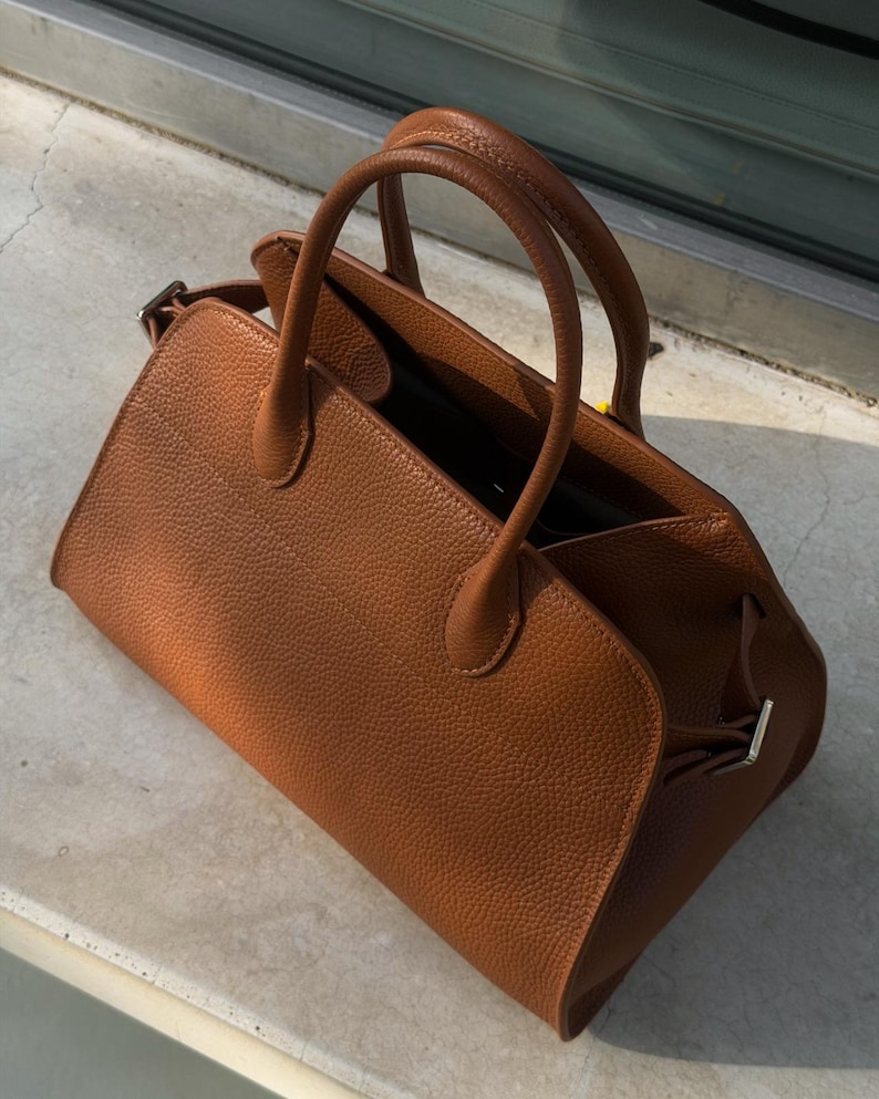 May include: A medium-sized, structured handbag in a rich cognac brown color with a distinct pebbled texture. The bag features two rounded top handles, a wide open-top design, and silver-toned metal hardware accents on the side attachments.