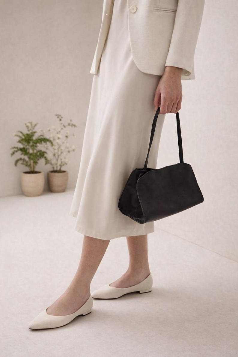 May include: A black handbag with a rectangular shape and curved top is held by a person. The person is wearing a cream-colored skirt, blazer, and ballet flats. Two small potted plants are in the background.
