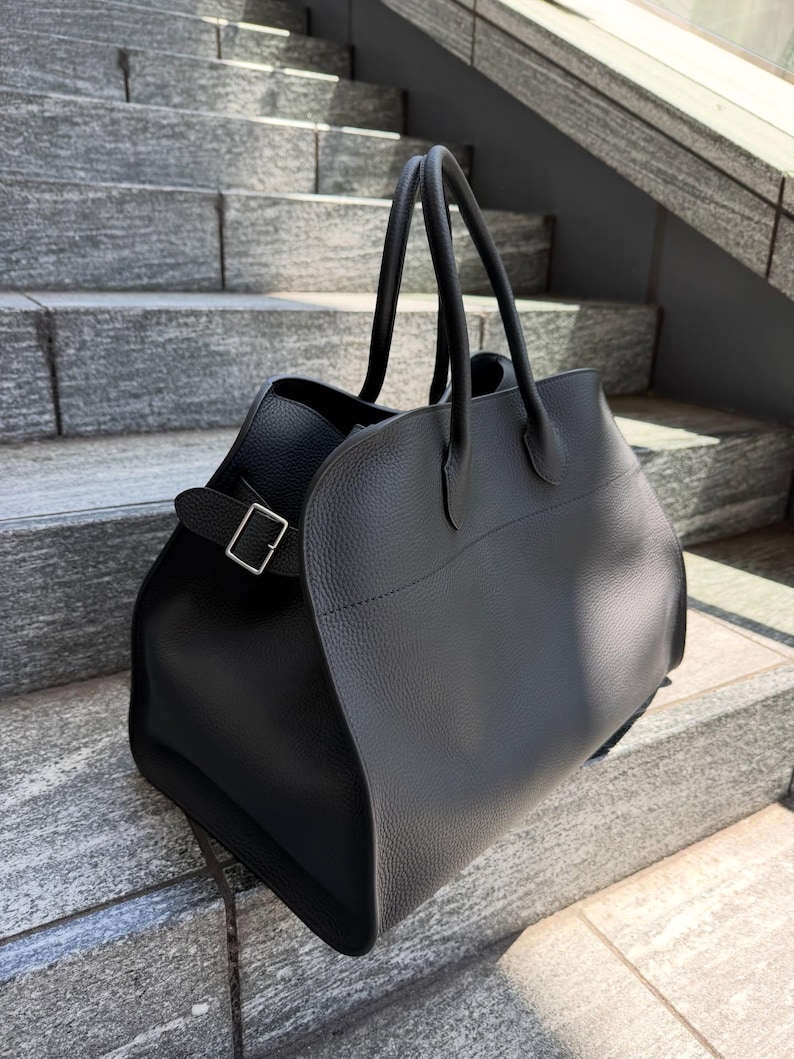 May include: A black textured leather handbag featuring two top handles and a side strap with a silver-toned metal buckle. The bag has a structured, wide-base design with a smooth, curved front panel and a pebbled finish throughout the exterior.