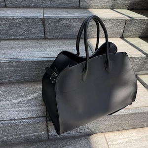 May include: A black handbag with a textured surface, featuring two rounded top handles and a side buckle detail. The bag has a structured base and a wide opening, showcasing a clean, solid color design with visible stitching along the top panel.