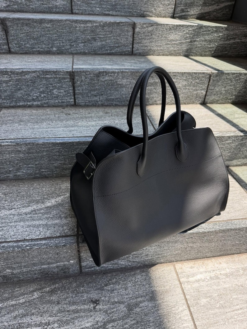 May include: A black handbag with a textured surface, featuring two rounded top handles and a side buckle detail. The bag has a structured base and a wide opening, showcasing a clean, solid color design with visible stitching along the top panel.