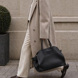 May include: A beige double-breasted wool coat worn over a white shirt and light-colored trousers, paired with dark brown leather loafers and a large black leather handbag featuring dual top handles and a side buckle detail.
