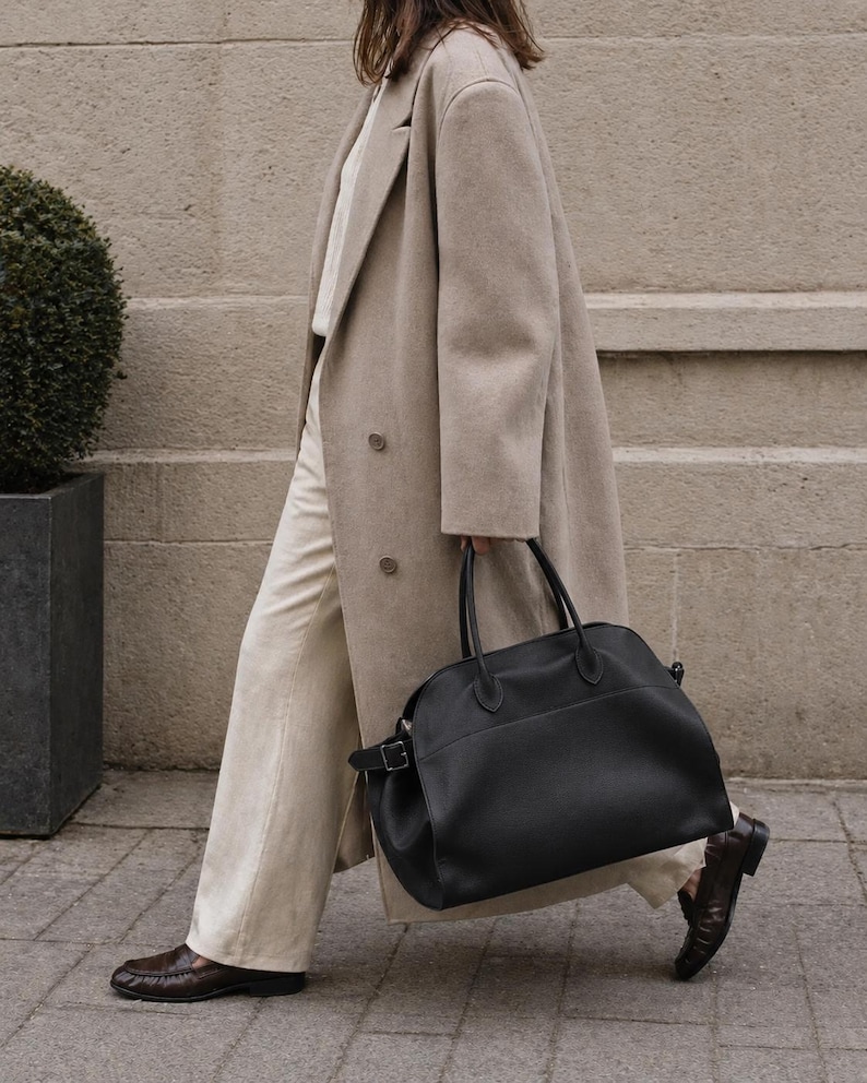 May include: A beige double-breasted wool coat worn over a white shirt and light-colored trousers, paired with dark brown leather loafers and a large black leather handbag featuring dual top handles and a side buckle detail.