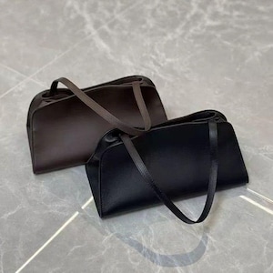 May include: Two leather shoulder bags. One is black, the other is brown. Both have a rectangular shape with curved edges and long, thin straps. The bags are placed on a light gray surface.