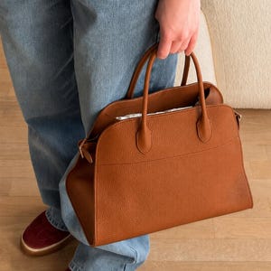 May include: A medium-sized, structured handbag in a rich cognac brown color, featuring a pebbled leather texture, two top handles, and a wide, open-top design with a curved silhouette. The bag has a horizontal seam detail across the front panel.