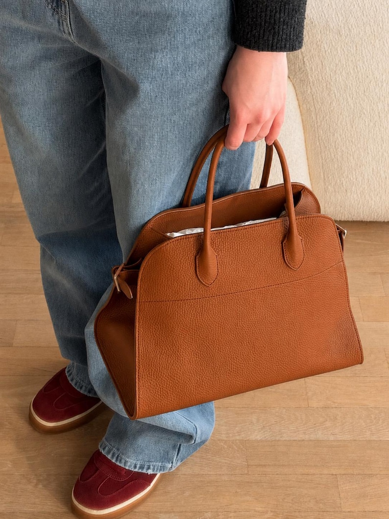 May include: A medium-sized, structured handbag in a rich cognac brown color, featuring a pebbled leather texture, two top handles, and a wide, open-top design with a curved silhouette. The bag has a horizontal seam detail across the front panel.