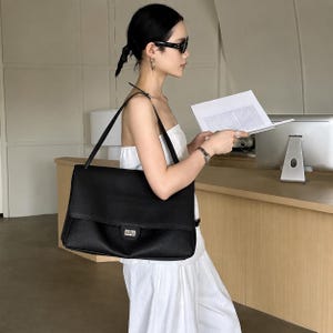 May include: A large black rectangular shoulder bag with a textured surface, a wide flap closure, and a silver-toned metal turn-lock clasp. The bag features a single shoulder strap and a structured silhouette, designed for carrying items on the shoulder.