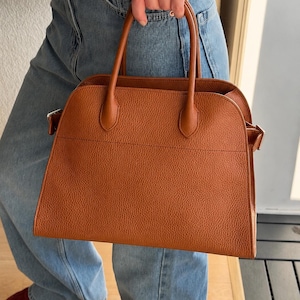 May include: A structured, cognac-colored handbag with a pebbled texture, featuring two top handles, a wide main compartment, and silver-toned metal hardware attachments on the sides for a strap, presented in a clean, minimalist design.