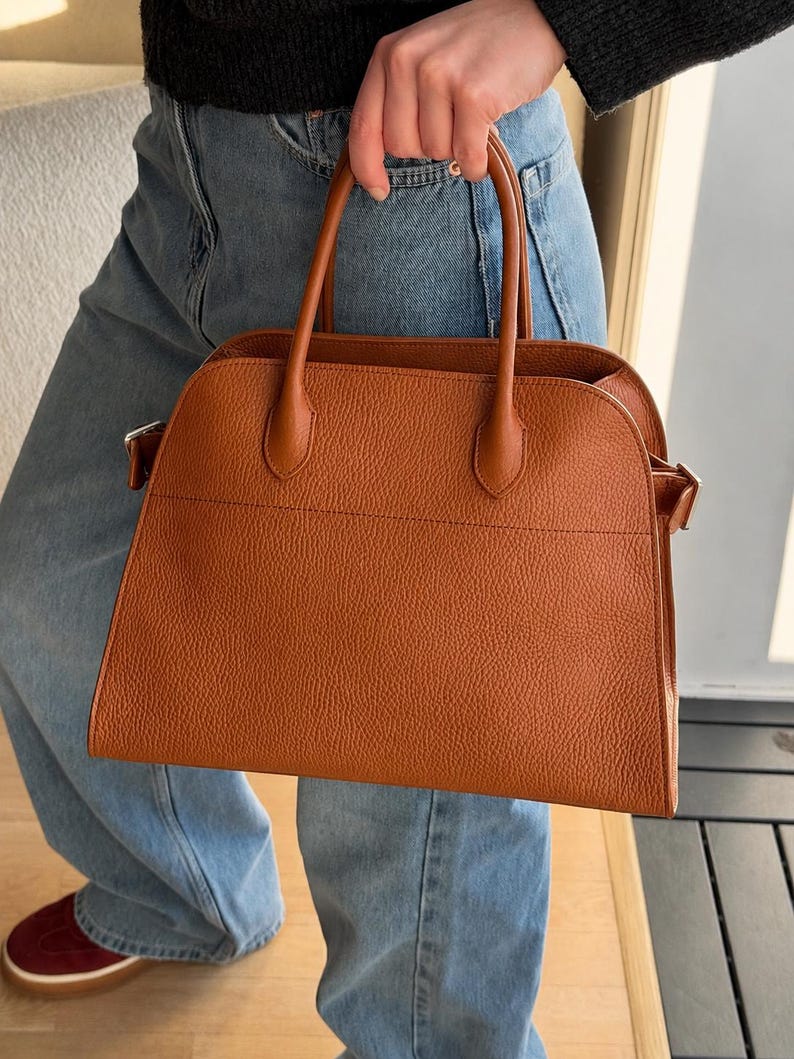 May include: A structured, cognac-colored handbag with a pebbled texture, featuring two top handles, a wide main compartment, and silver-toned metal hardware attachments on the sides for a strap, presented in a clean, minimalist design.
