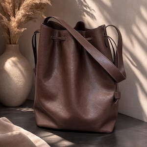 May include: A brown leather bucket bag with a shoulder strap. The bag has a drawstring closure and is sitting on a dark surface. A vase with dried plants is in the background.