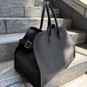 May include: A black textured leather handbag featuring two top handles and a side strap with a silver-toned metal buckle. The bag has a structured, wide-base design with a smooth, curved front panel and a pebbled finish throughout the exterior.