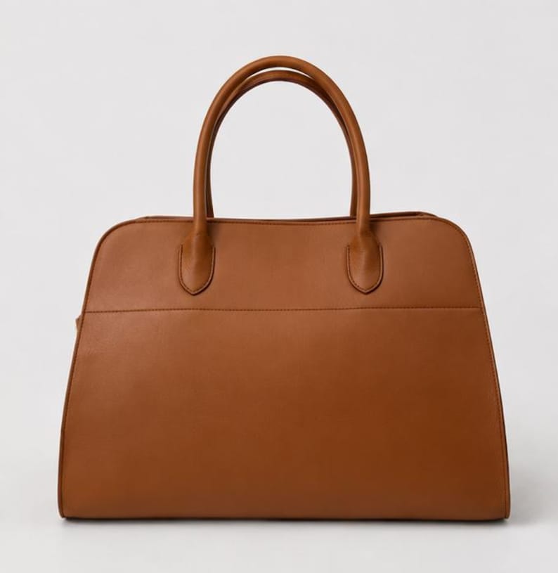 May include: A structured tan handbag with a smooth finish, featuring two rounded top handles attached to the main body with reinforced stitching. The bag has a wide, flat base and a horizontal seam detail across the front panel of the exterior.