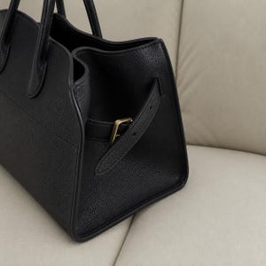 May include: A black handbag with a textured surface, featuring two top handles and a side strap secured with a gold-toned metal buckle. The bag has a structured design with visible stitching along the edges and a smooth interior lining visible.