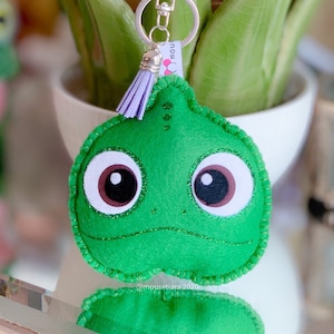 May include: A green felt Pascal keychain with a silver clasp and a purple tassel. The keychain features a cartoon-style design with large brown and white eyes, and a glitter outline. The background includes a green plant in a white pot.
