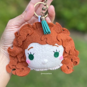 Puede incluir: Un llavero de fieltro con un personaje de dibujos animados con cabello rojo rizado, ojos verdes y una cara blanca. El llavero tiene un borla verde y un llavero de metal dorado. El texto "@mousetiara" es visible en el llavero.