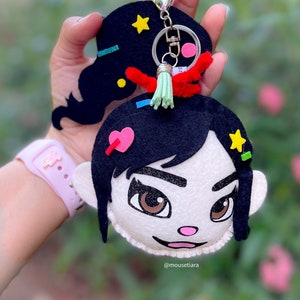 Puede incluir: Un llavero de fieltro con un personaje de dibujos animados con cabello negro, ojos marrones y una nariz rosa. El personaje lleva una camisa blanca con un corazón rosa y una estrella amarilla. El llavero está unido a un llavero plateado con una borla verde.