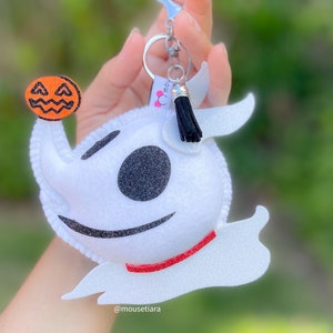 Può includere: Portachiavi in feltro bianco con un viso bianco e nero, un colletto rosso e un fiocco nero. Il portachiavi tiene una piccola zucca in feltro arancione con un viso nero.