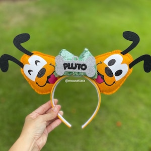Puede incluir: Una diadema de fieltro amarilla con dos orejas de Pluto. Las orejas tienen la forma de la cabeza de Pluto con orejas negras y ojos blancos. Un lazo verde y plateado con una etiqueta en forma de hueso que dice "PLUTO" está en el centro de la diadema.