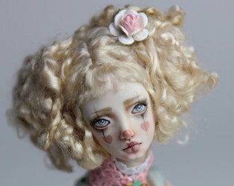 May include: A porcelain doll with blonde curly hair, wearing a white and blue dress with a pink lace collar and a flower in her hair. The doll has painted-on makeup, including red hearts on her cheeks.