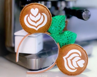 May include: Brown felt mouse ears with a white latte art design on each ear. The ears are attached to a white headband with a green sparkly bow on one side. The ears are in front of a silver espresso machine.