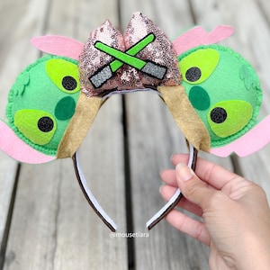 May include: Green and pink felt character ears with a brown felt headband. The character ears feature a pink sparkly bow with green lightsabers crossed on top. The headband has the text "@mousetiara" on it.