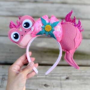 May include: Pink and teal mouse ears with a pink bow and a flower with a yellow center. The ears are shaped like a dragon with a pink tail and a pink head with large eyes. The headband is white.