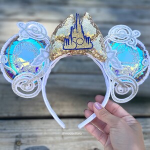 May include: White and gold Minnie Mouse ears with a blue and white iridescent design and a gold castle with the number 50 on it.