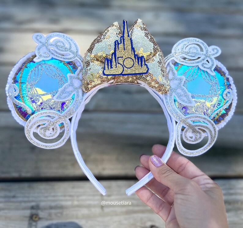 May include: White and gold Minnie Mouse ears with a blue and white iridescent design and a gold castle with the number 50 on it.