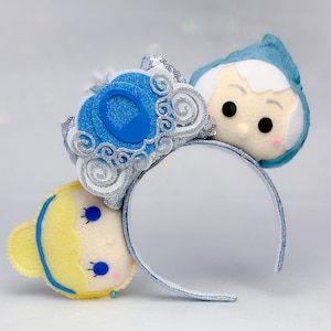 May include: A pair of blue and white Minnie Mouse ears with a blue and silver glitter carriage charm and a yellow and white felt Cinderella character on one ear and a blue felt character on the other ear.