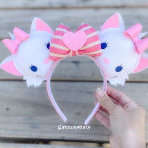 Può includere: Fascia per capelli con orecchie di gatto in feltro bianco e un fiocco a righe rosa e bianche con un cuore rosa al centro. La fascia è decorata con occhi blu e dettagli rosa.