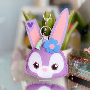 May include: Purple felt bunny keychain with a pink flower, green tassel, and a silver key ring. The bunny has a white face with black eyes and a pink nose. The keychain has a small purple Mickey Mouse ear on the back.