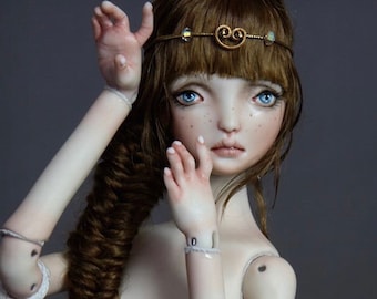 May include: A porcelain doll with long brown hair styled in a braid, wearing a gold headband with a decorative design. The doll has blue eyes and a delicate facial expression.