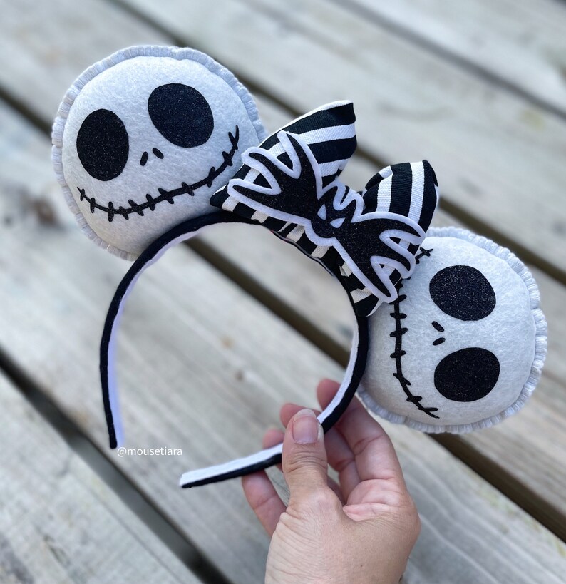 May include: Black and white felt headband with a black and white striped bow and two felt ears shaped like Jack Skellington's face from the Nightmare Before Christmas. The headband has a black and white striped bow with a black bat design in the center. The ears are white with black eyes, a black stitched mouth, and black glitter details.