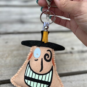 May include: A felt keychain shaped like a character with a black hat, one blue eye, and a wide, toothy grin. The keychain has a silver key ring and a yellow tassel.